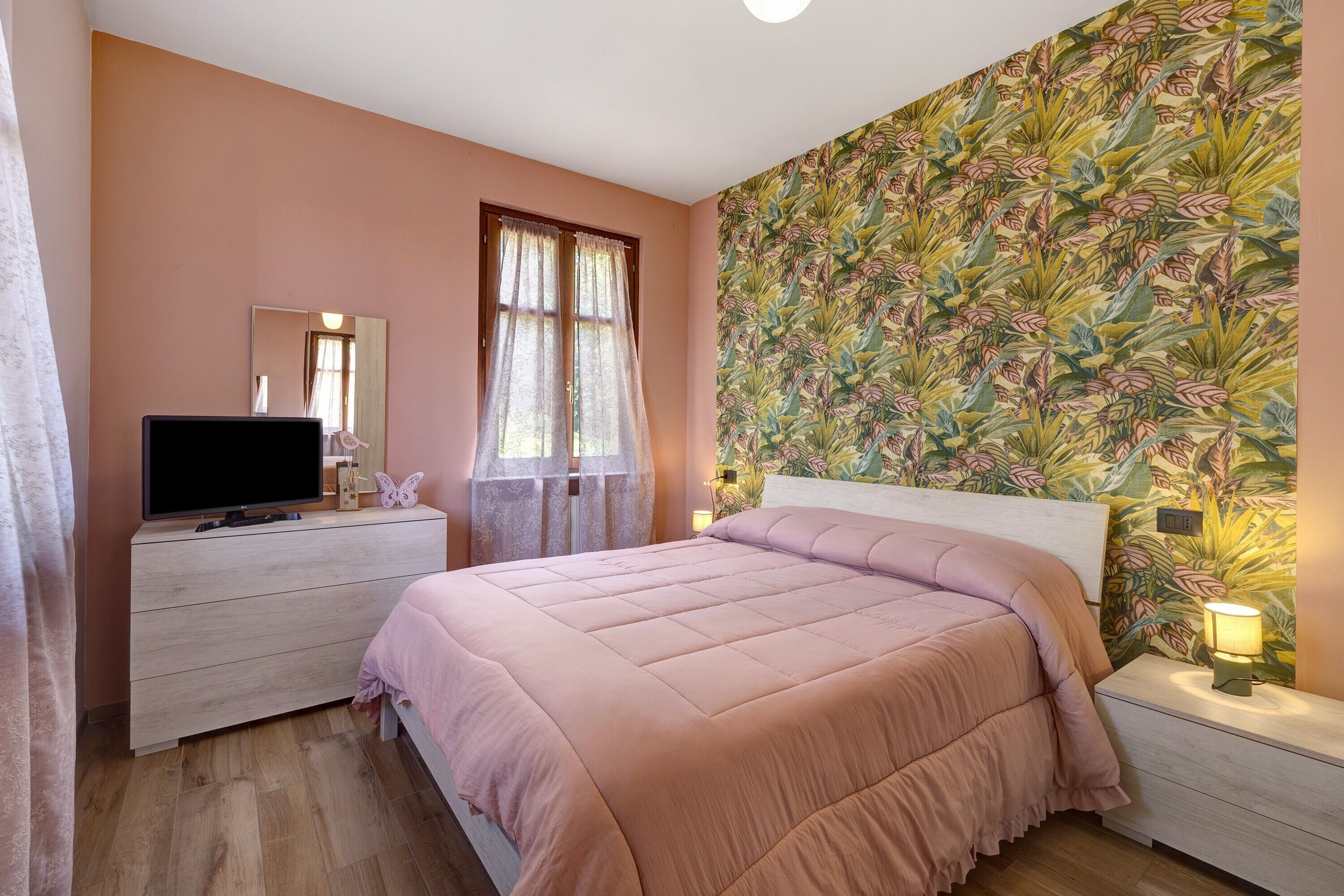 A bedroom with a bed, a dresser, a television, a window with curtains, and a floral wallpaper.