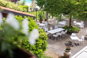 Une terrasse extérieure avec des tables et des chaises blanches, entourée de verdure et bordée d’un chemin pavé.