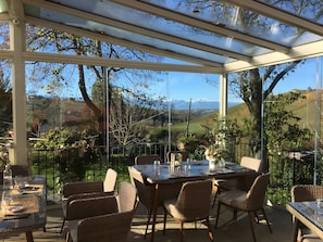 A covered outdoor dining area with a table and chairs, large windows, and a view of the countryside.
