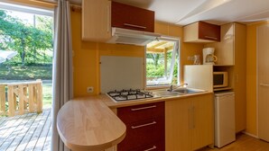 A compact kitchen with a gas stove, sink, and microwave, wooden cabinets, and a view of the outdoors. - 3★ Campsite in Périgord Noir – Mobile Home with Pool, Fishing & Family Activities (Saint-Martial-de-Nabirat)