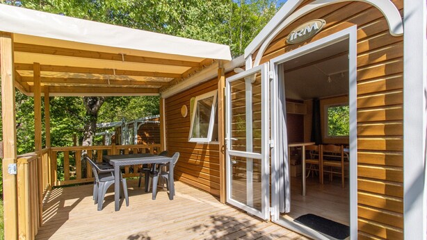 A wooden deck with a table and chairs, a covered area, and a building entrance with a sign. - 3★ Campsite in Périgord Noir – Mobile Home with Pool, Fishing & Family Activities (Saint-Martial-de-Nabirat)