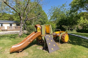 Une aire de jeux avec un toboggan et une rampe, entourée d’arbres et bordée d’un sentier.