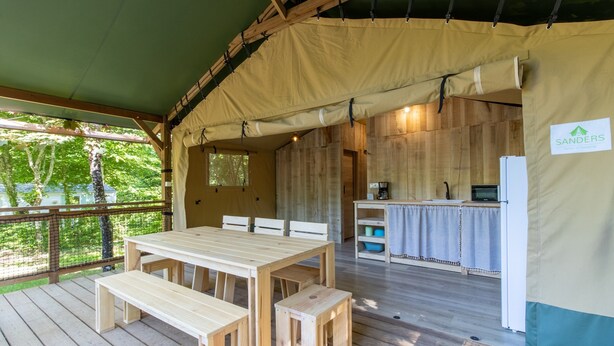 Una terraza de madera con una mesa y bancos, una zona de cocina con refrigerador, y un cartel que dice 'SANDERS'.