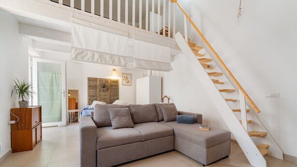A modern living room with a grey sectional sofa, a wooden staircase, and a view of the dining area. - Holiday Home 'Sa Nue' with Private Terrace, Wi-Fi and Air Conditioning (Calasetta)