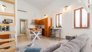A modern living room with a grey sofa, a wooden dining table with chairs, a kitchen area with wooden cabinets, and a small balcony with a potted plant. - Holiday Home 'Sa Nue' with Private Terrace, Wi-Fi and Air Conditioning (Calasetta)