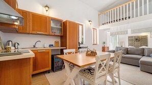 A modern kitchen with wooden cabinets, a dining area with a wooden table and white chairs, and a living room with a grey sofa and a white railing. - Holiday Home 'Sa Nue' with Private Terrace, Wi-Fi and Air Conditioning (Calasetta)