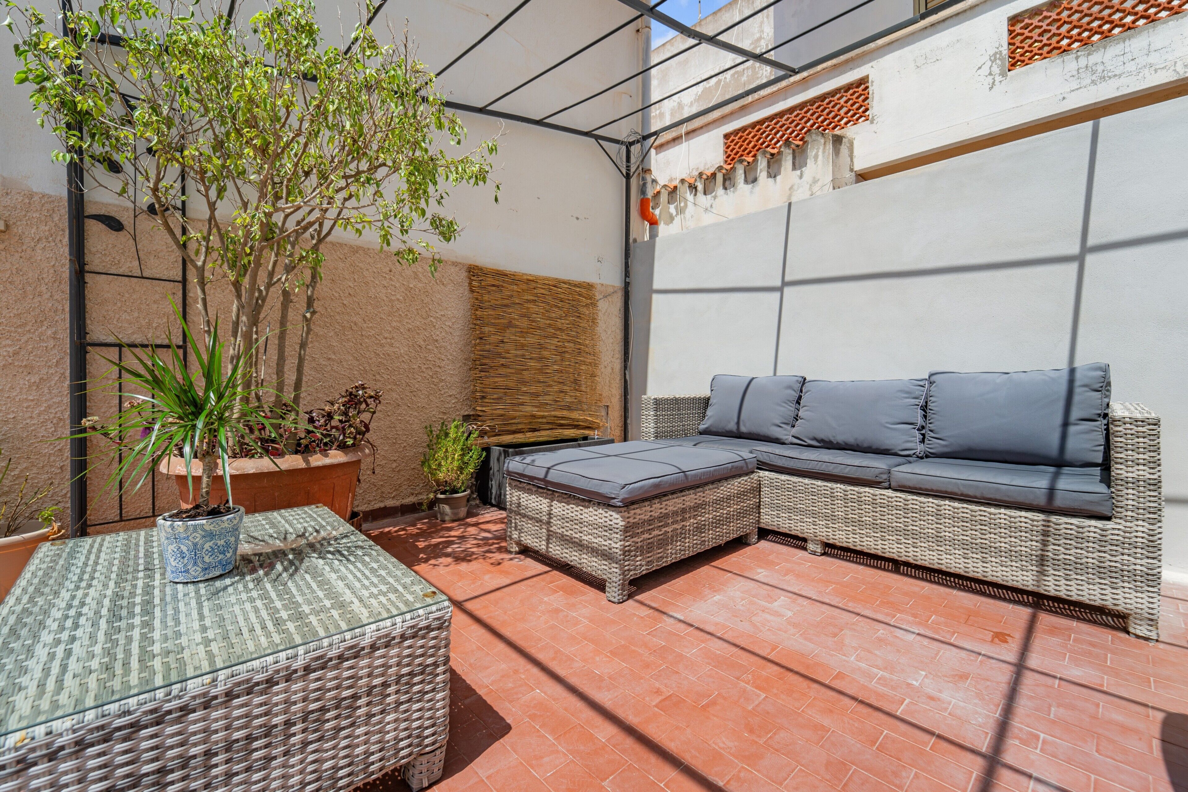 Eine überdachte Terrasse mit einem Rattan-Sofa, einem kleinen Tisch und Kübelpflanzen.
