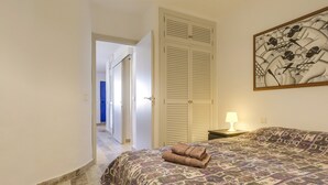 Een slaapkamer met een bed met een decoratieve spreidek, een opgerolde handdoek op het bed, een nachtkastje met een lamp en een groot kunstwerk in een lijst aan de muur.