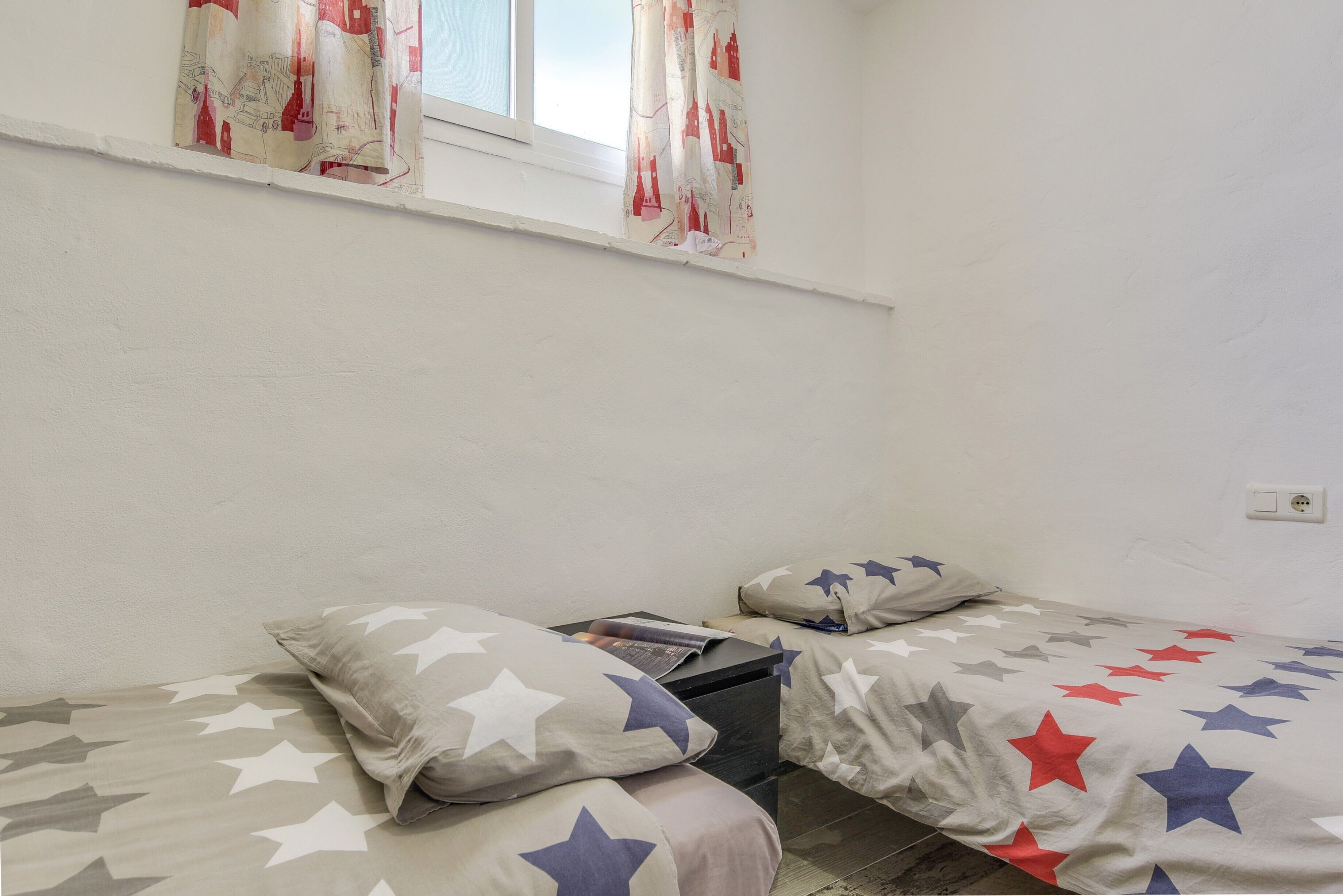 Habitación con dos camas, cada una con ropa de cama estampada con motivos de estrellas, una ventana con cortinas y un enchufe visible.