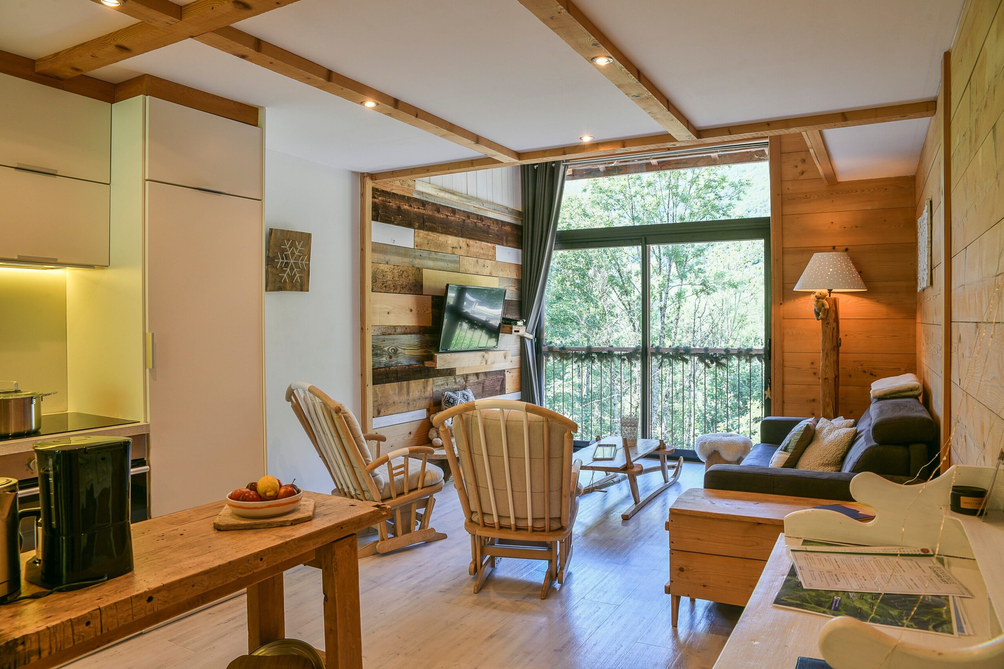 A wooden interior with a dining table, chairs, a sofa, and a large window with a view of trees.