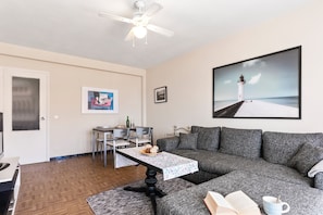 A living room with a sofa, a table with a book and a mug, a ceiling fan, and a painting of a lighthouse.