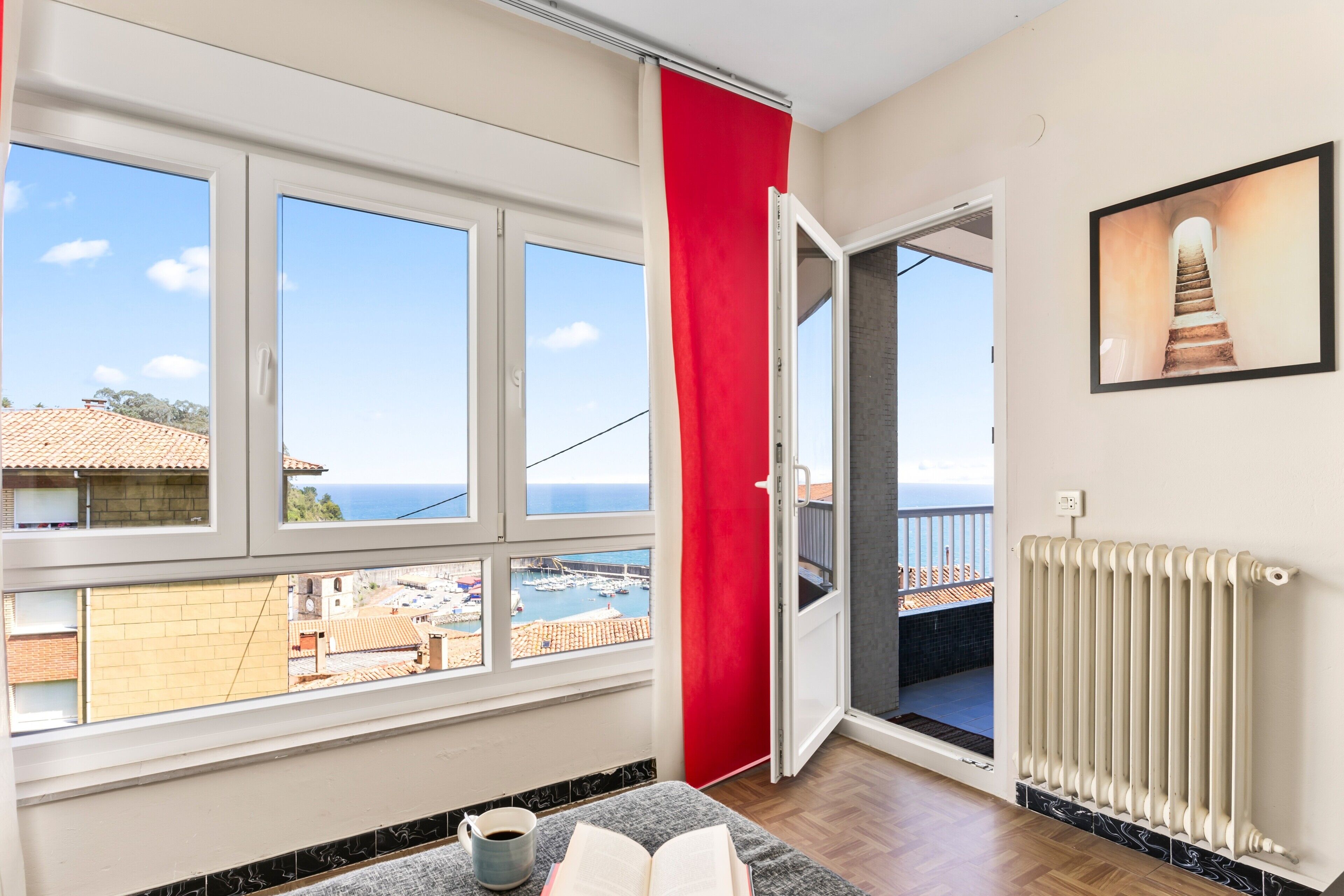 A room with a large window, a red curtain, a radiator, and a view of the sea and buildings.