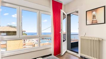 A room with a large window, a red curtain, a radiator, and a view of the sea and buildings.