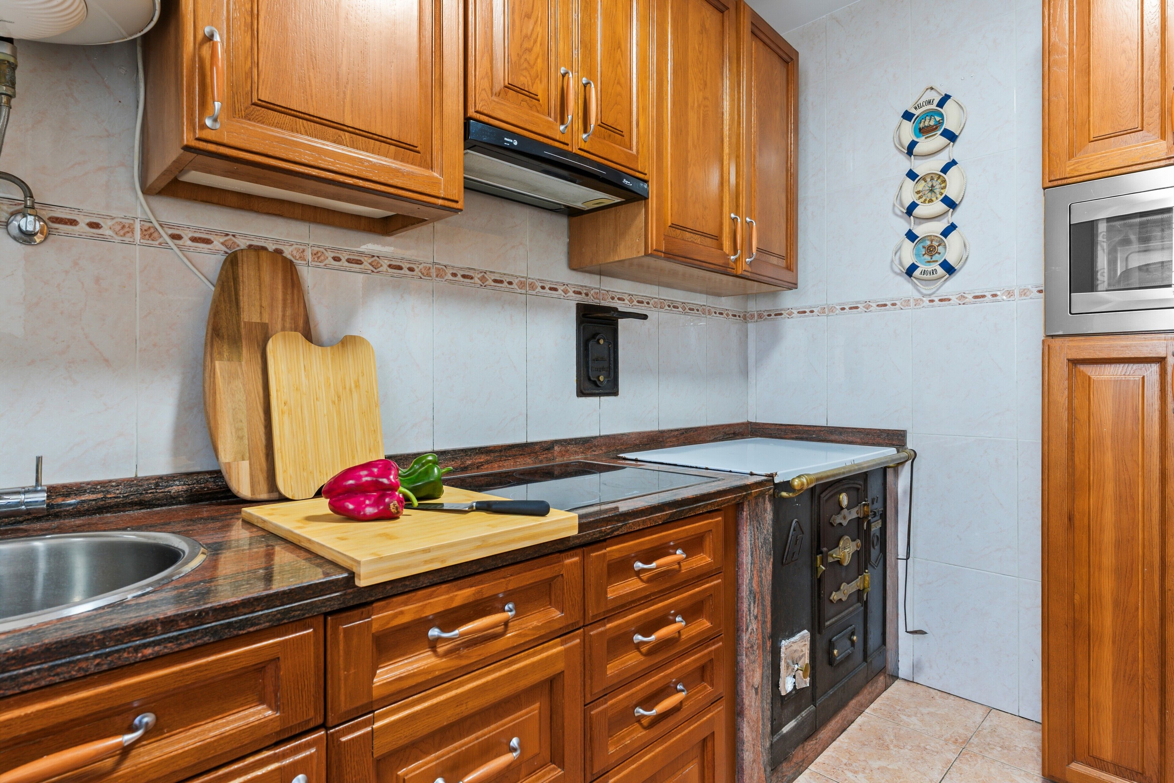 Uma cozinha com armários de madeira, pia de aço inoxidável, tábua de corte com vegetais e micro-ondas.