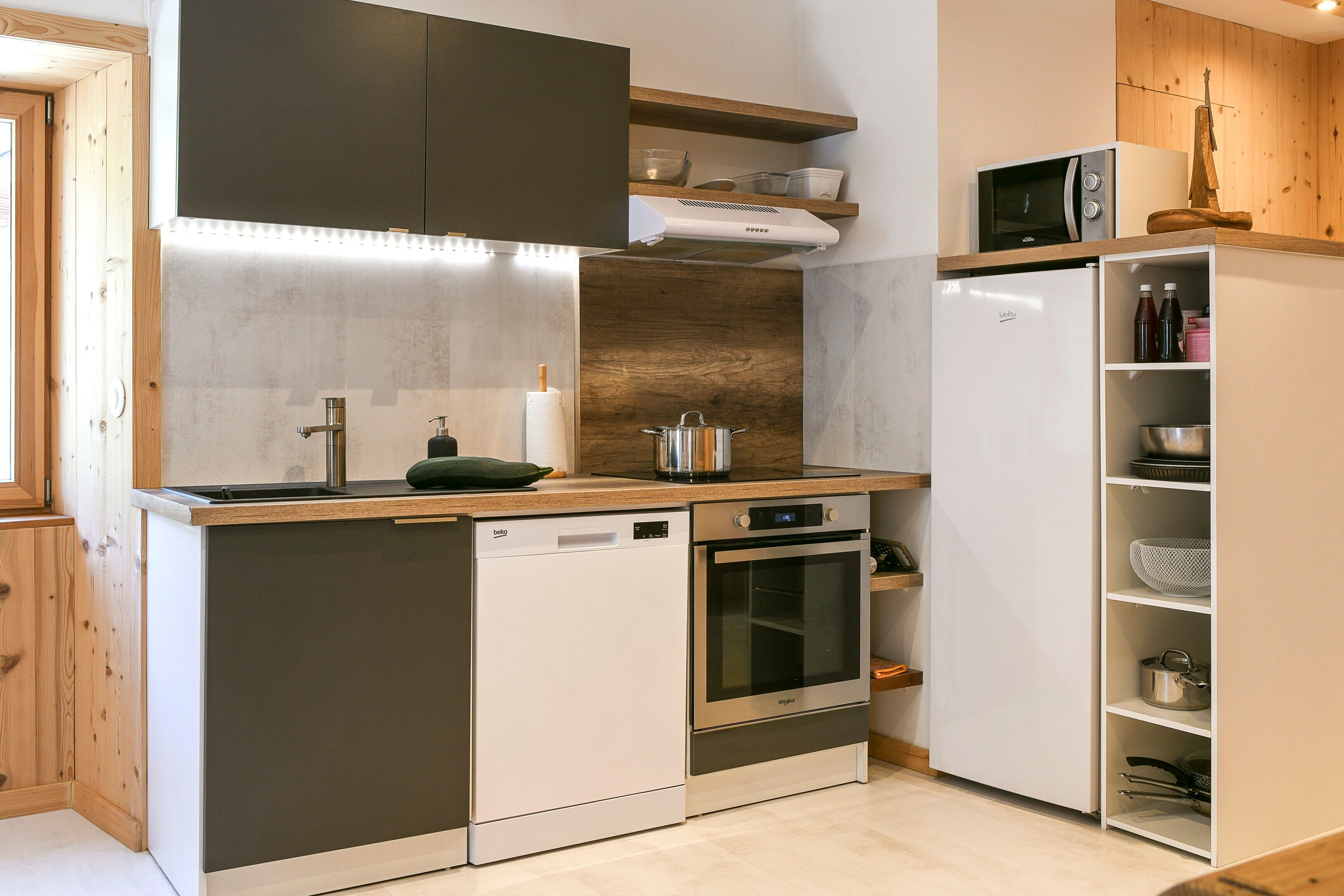 A compact kitchen with a dishwasher, oven, microwave, and wooden shelves.