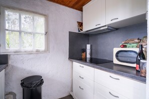 A kitchen with white cabinets, a microwave, a trash bin, and a window with lace curtains. - Holiday Home 'Le Refuge Marin' (Le Pradet)
