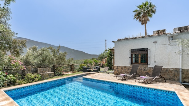 A swimming pool with lounge chairs and a stone wall, surrounded by greenery and a mountain view.