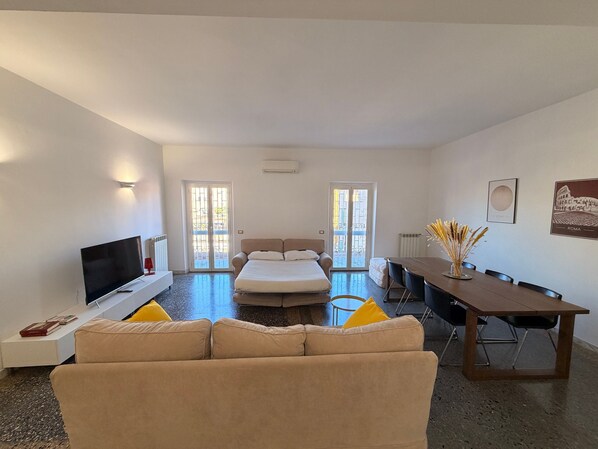 A modern hotel room with a sofa, a bed, a TV, a dining table, and two doors leading to a balcony.