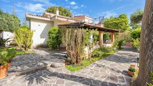 A stone-paved courtyard with a white building, a pergola, and various potted plants.