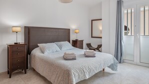 Een slaapkamer met een bed, nachtkastjes, lampen, een spiegel en uitzicht door een raam met gordijnen.