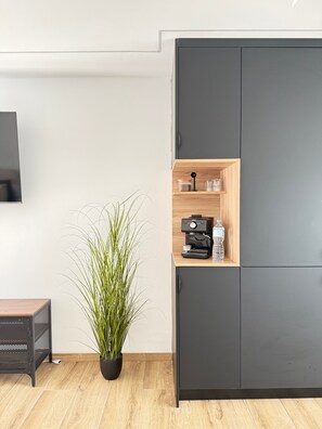 Una habitación moderna con un armario gris oscuro, un estante de madera con una cafetera, una planta en maceta y una pequeña mesa.