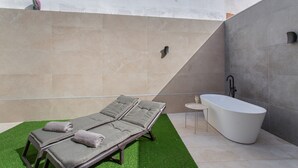 A modern bathroom with a freestanding bathtub, two sun loungers with towels, and a small table with a glass.