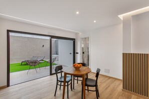 Un espace de repas moderne avec une table et des chaises en bois, un saladier rempli de fruits et une vue sur un patio extérieur avec une pelouse verdoyante et des sièges.