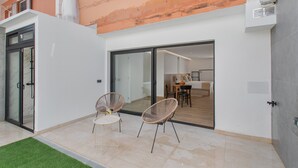 A modern patio with two wicker chairs, a glass door leading to an interior dining area, and a white exterior wall.