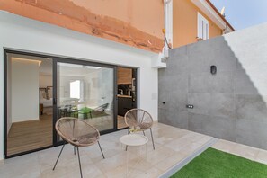 Un patio exterior moderno con dos sillas de mimbre y una mesita, una puerta corrediza de vidrio que da a un espacio interior, y una pared de baldosas grises.