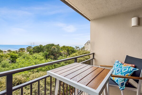 A balcony with a wooden table and a chair, overlooking a beach and greenery.