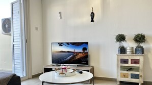 En stue med et fladskærms-tv på et stativ, et rundt bord med bakke, et hvidt skab med pynteting og en skydedør i glas.
