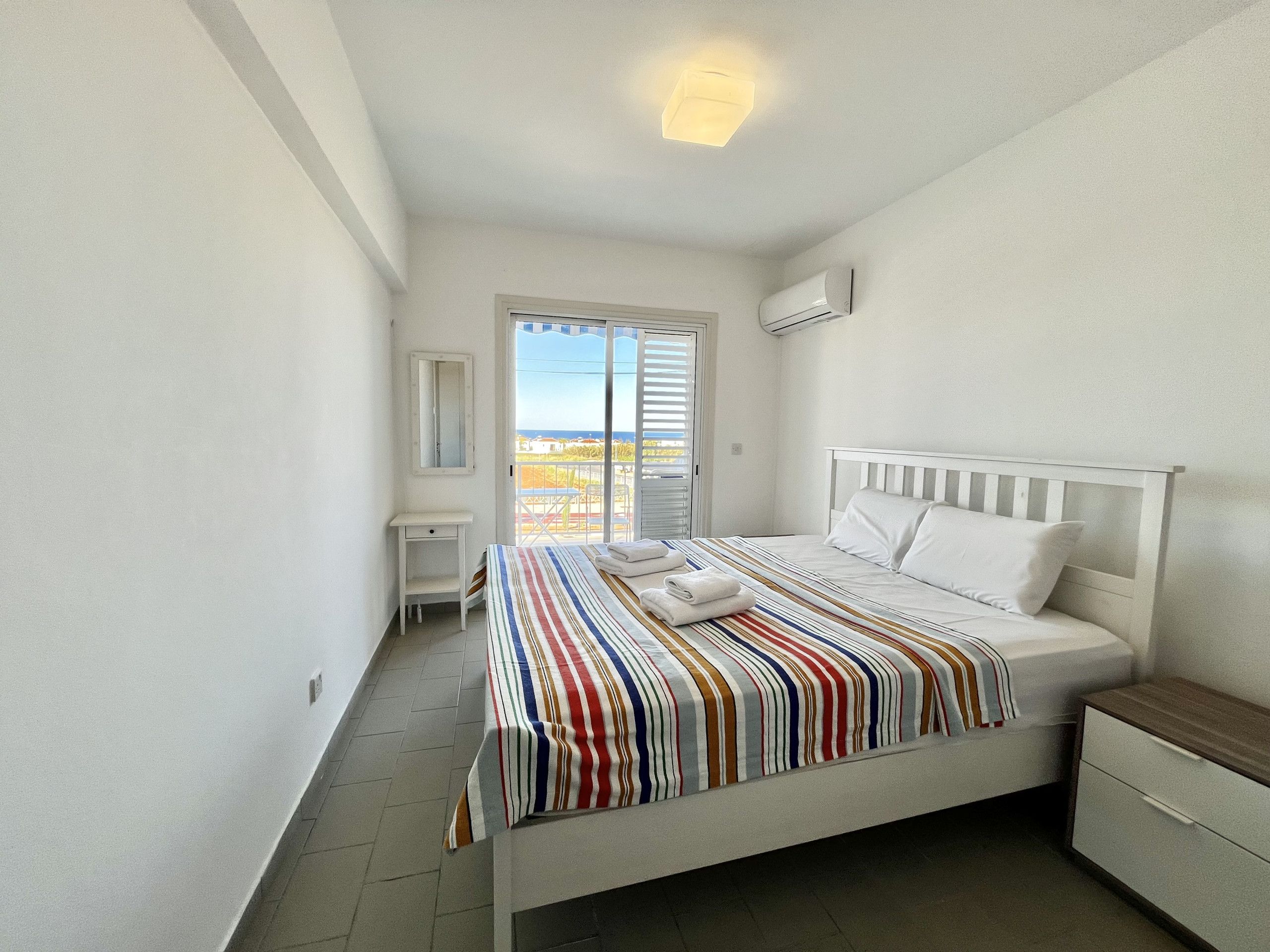 A bedroom with a bed, a bedside table, a nightstand, a mirror, and a sliding glass door leading to a balcony.