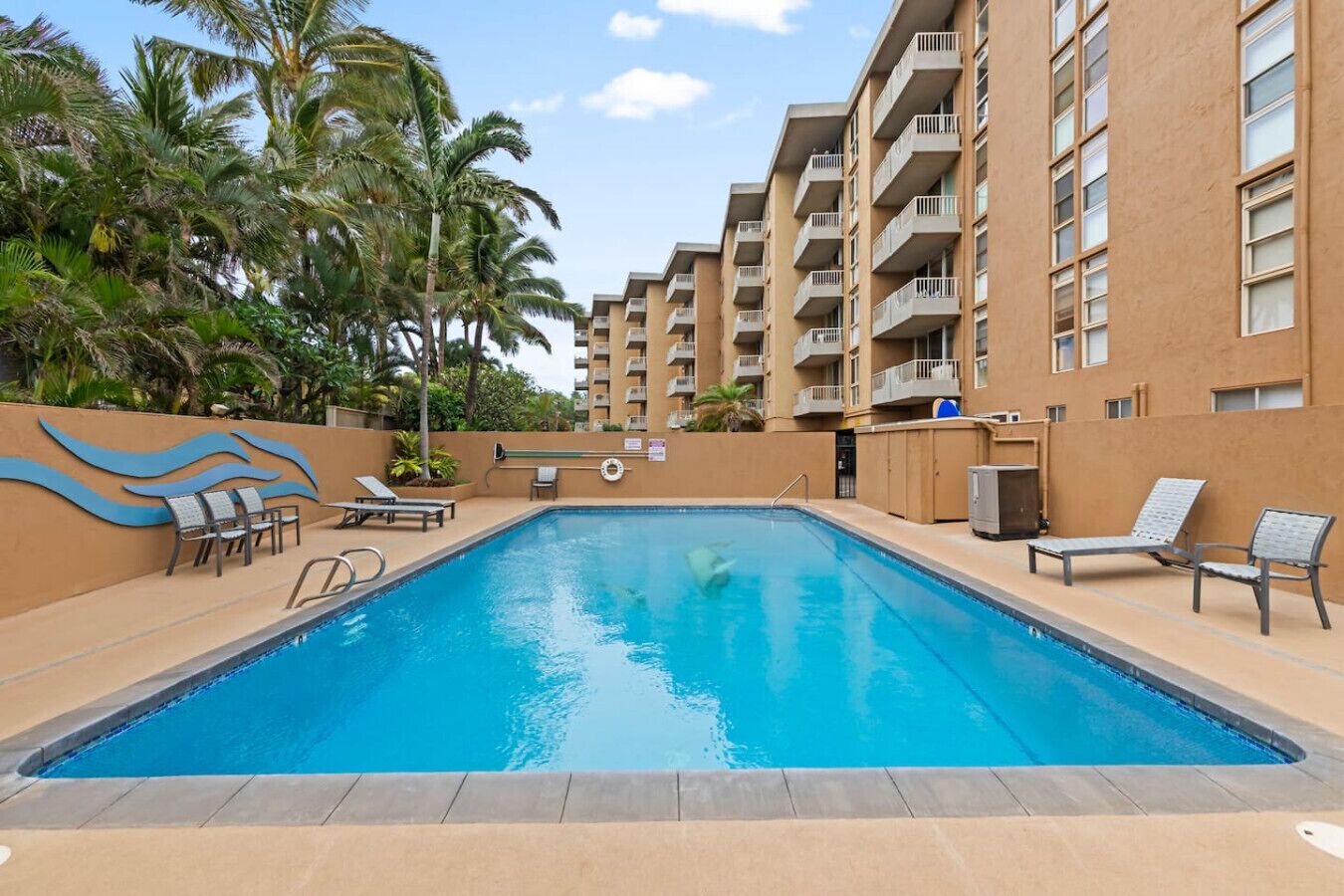 A swimming pool with lounge chairs and palm trees, surrounded by a building with balconies.