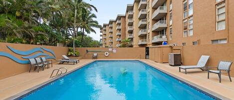 A swimming pool with lounge chairs and palm trees, surrounded by a building with balconies.