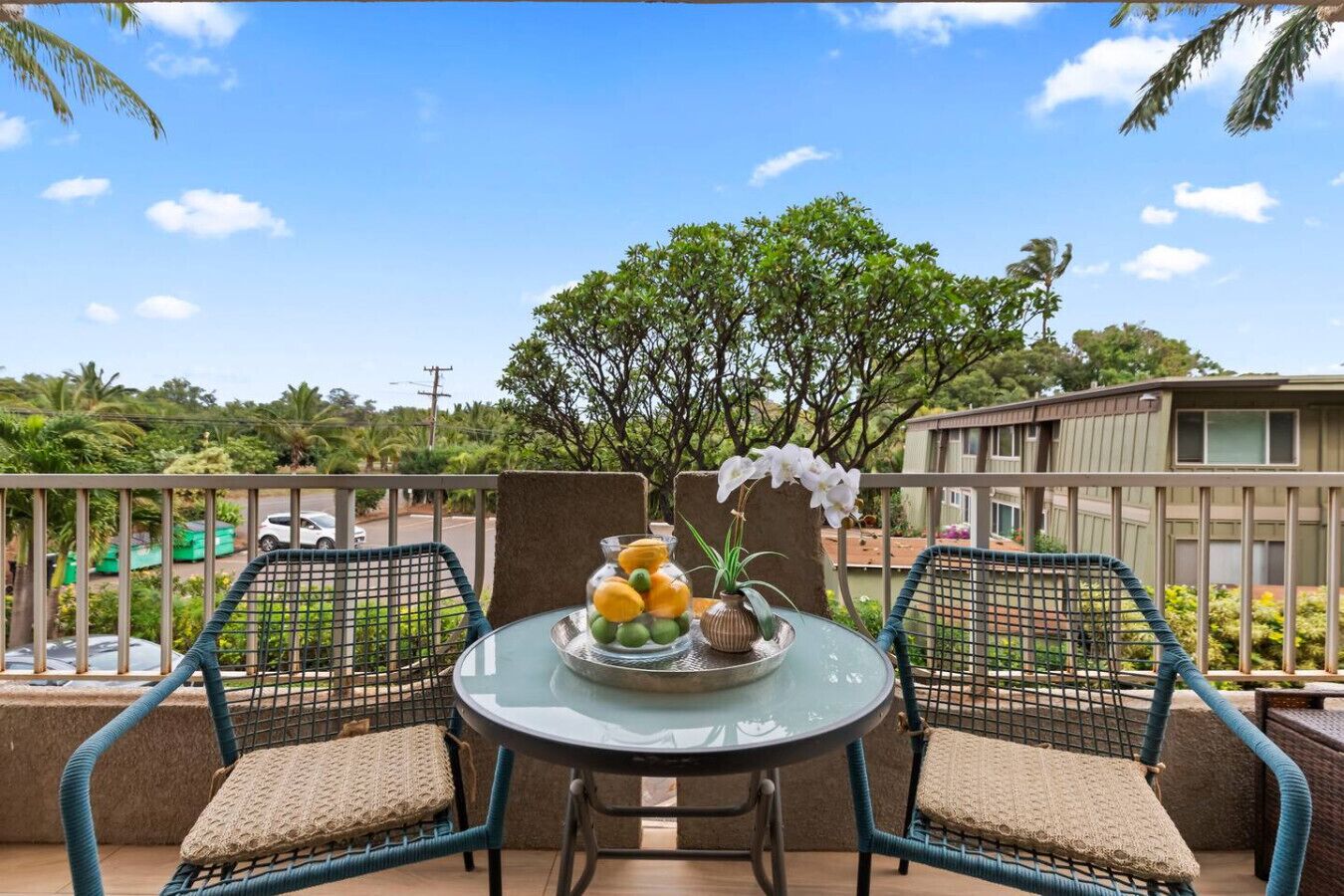 A balcony with two wicker chairs, a glass table with fruit and flowers, and a view of a residential area with trees and buildings.