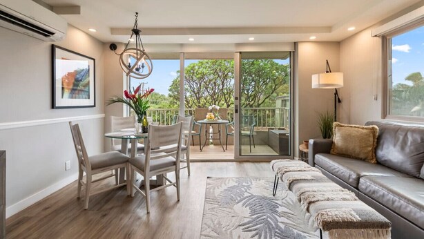 A modern living room with a sofa, a dining table with chairs, a chandelier, and a balcony with a view of trees.