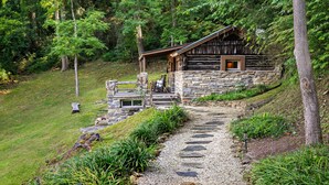 A rustic cabin with a stone foundation, a wooden deck, and a gravel path leading to the entrance.