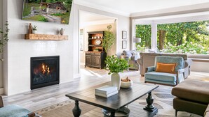 A modern living room with a fireplace, a large TV mounted on the wall, and a view of the outdoors through large windows.