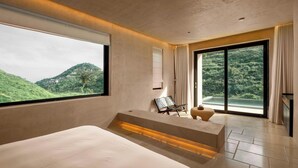 A modern bedroom with a large bed, a bench, a chair, and a view of a green landscape through large windows.
