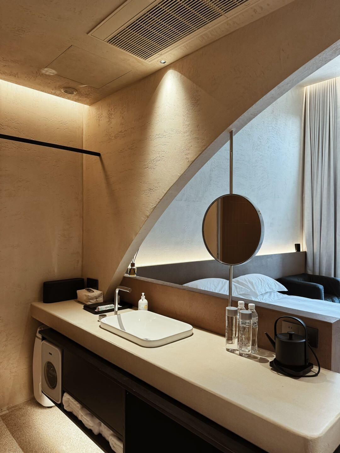 A modern hotel bathroom with a curved vanity, a large mirror, a sink, and a coffee machine.