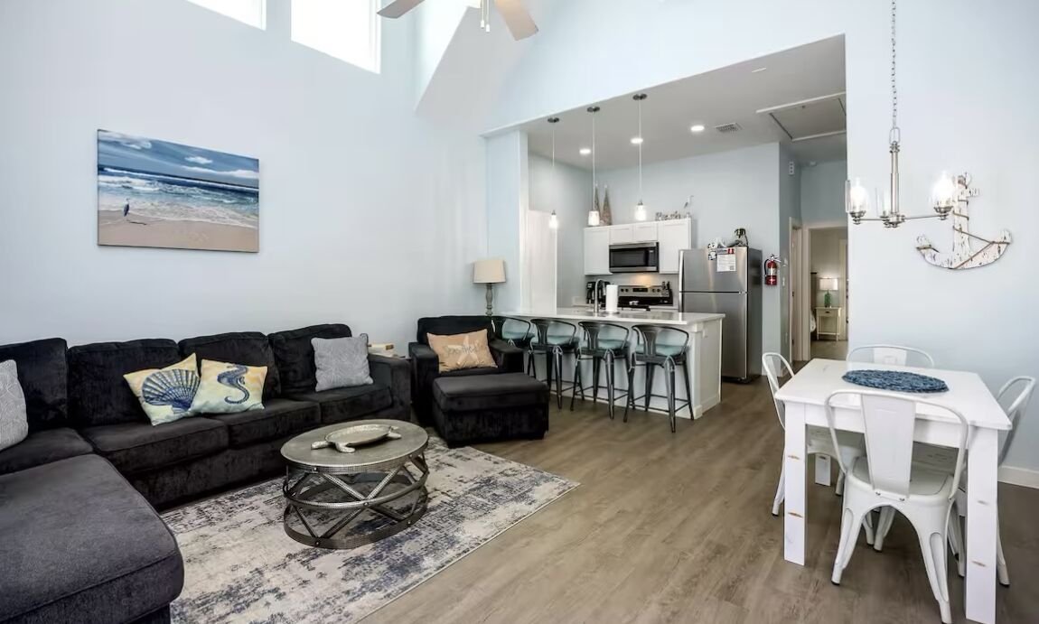 A modern living room with a sofa, coffee table, and a dining area with a white table and chairs. A kitchen with a bar area is visible in the background.