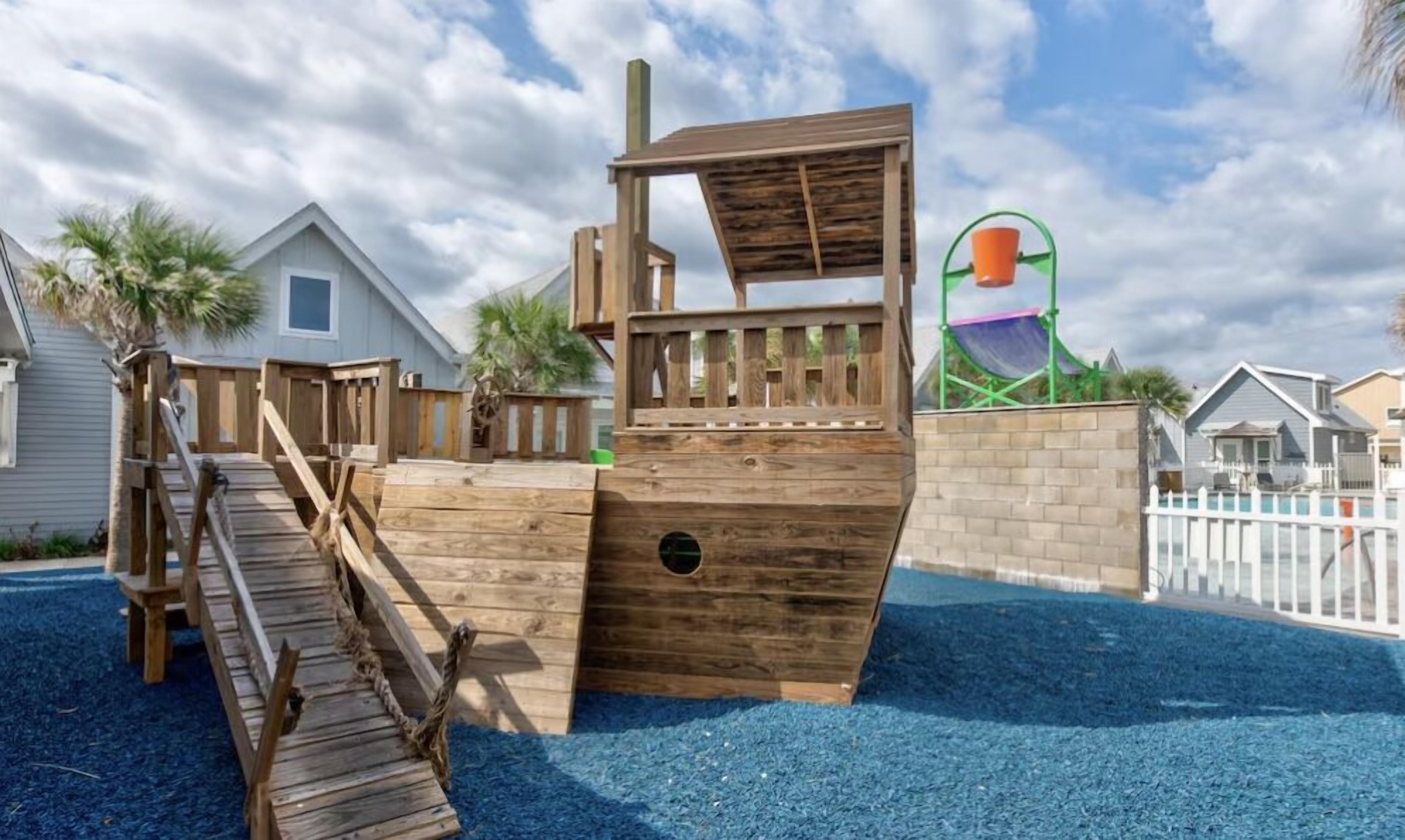 Une structure de jeu en bois, dotée d’un toboggan et d’un mur d’escalade, entourée d’une clôture en bois peinte en blanc et d’une surface de jeu bleue.