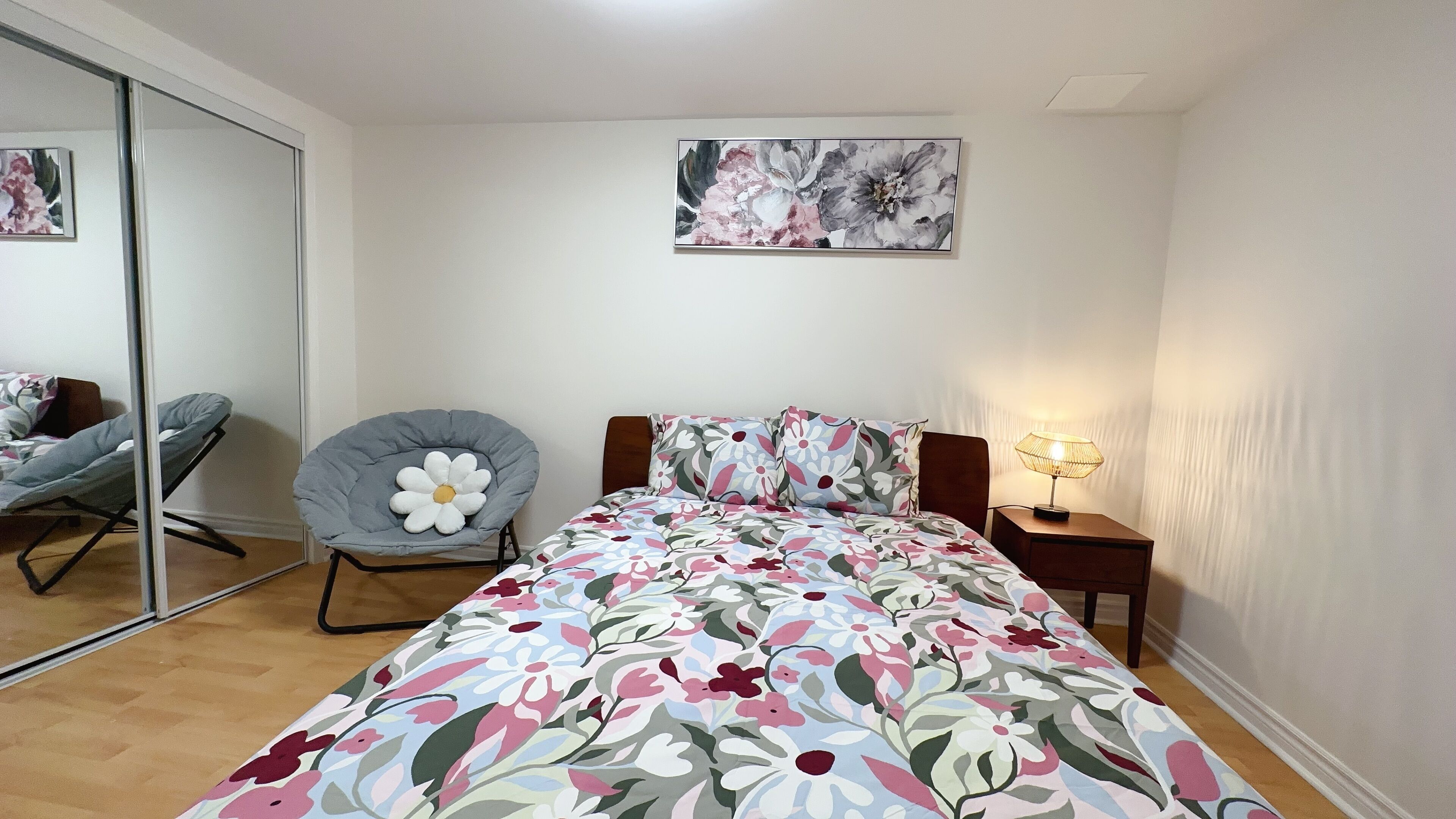 Une chambre à coucher comprenant un lit, une chaise, une table de chevet avec une lampe, un miroir et un tableau représentant des fleurs accroché au mur.
