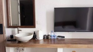 A bathroom with a wooden vanity, a large mirror, a white sink, a black TV mounted on the wall, and two bottles of water.