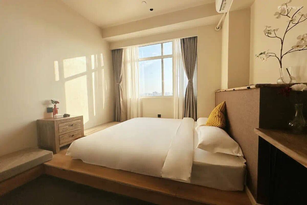 Una camera da letto con un letto, un comodino, una cassettiera, una finestra con tende e un vaso con fiori sulla cassettiera.