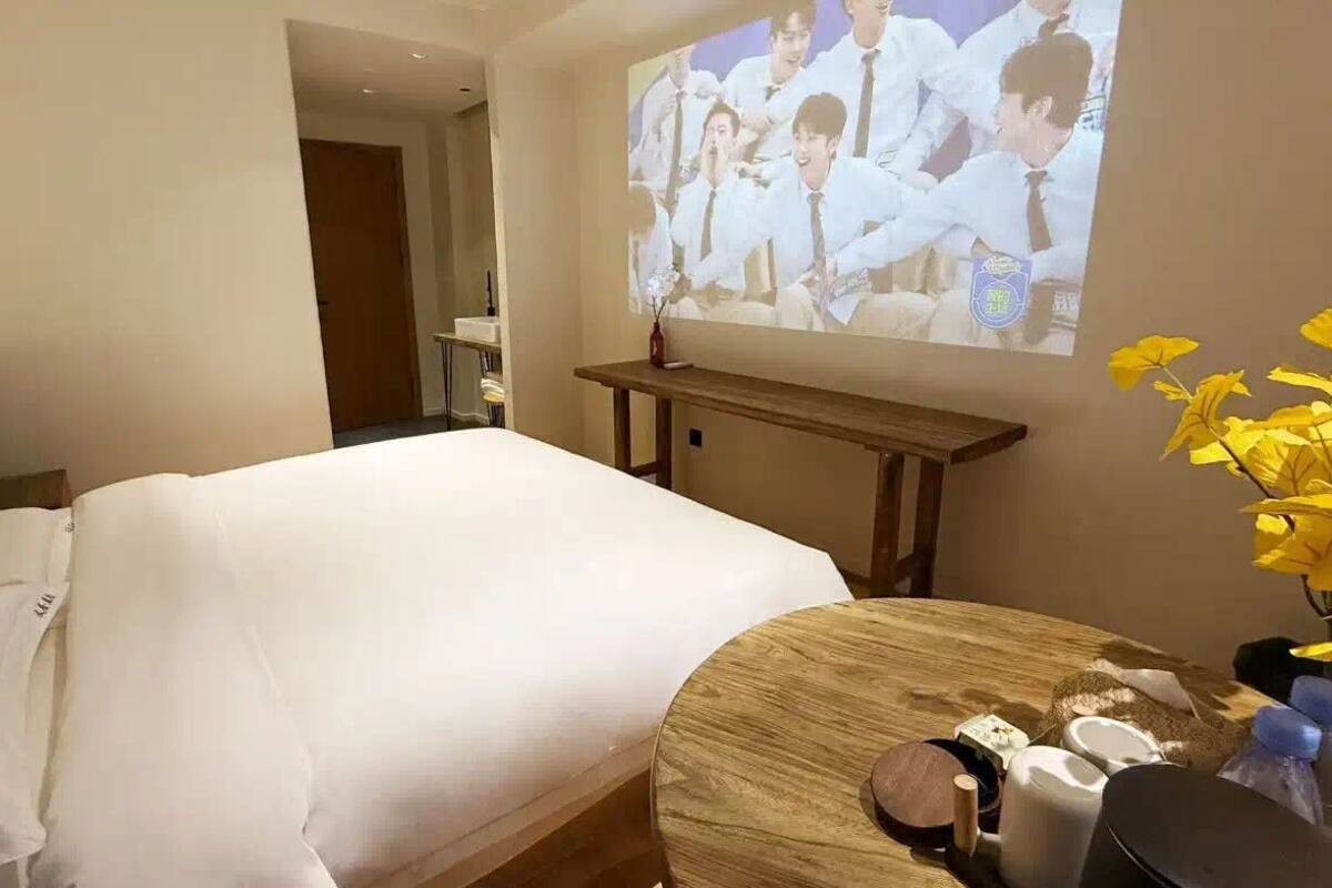 Una camera d'albergo con un letto, un comodino in legno con articoli da toilette e un televisore a parete che mostra un gruppo di persone.