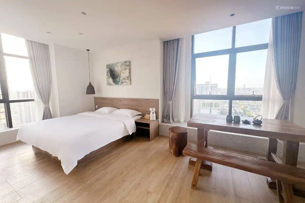 Una camera d'albergo moderna con un letto grande, una panca di legno, un tavolino e vista sulla città attraverso ampie finestre.
