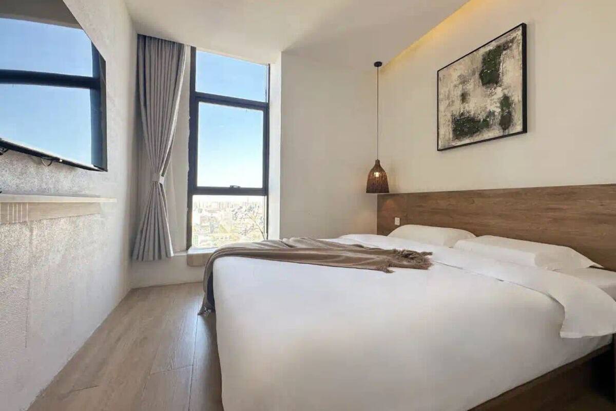 Una moderna camera d'albergo con un ampio letto, una testiera in legno, una finestra con tende, un'opera d'arte appesa al muro e una lampada a sospensione.