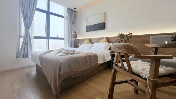 A modern hotel room with a large bed, a wooden nightstand, a small table, and a view of the city through large windows.