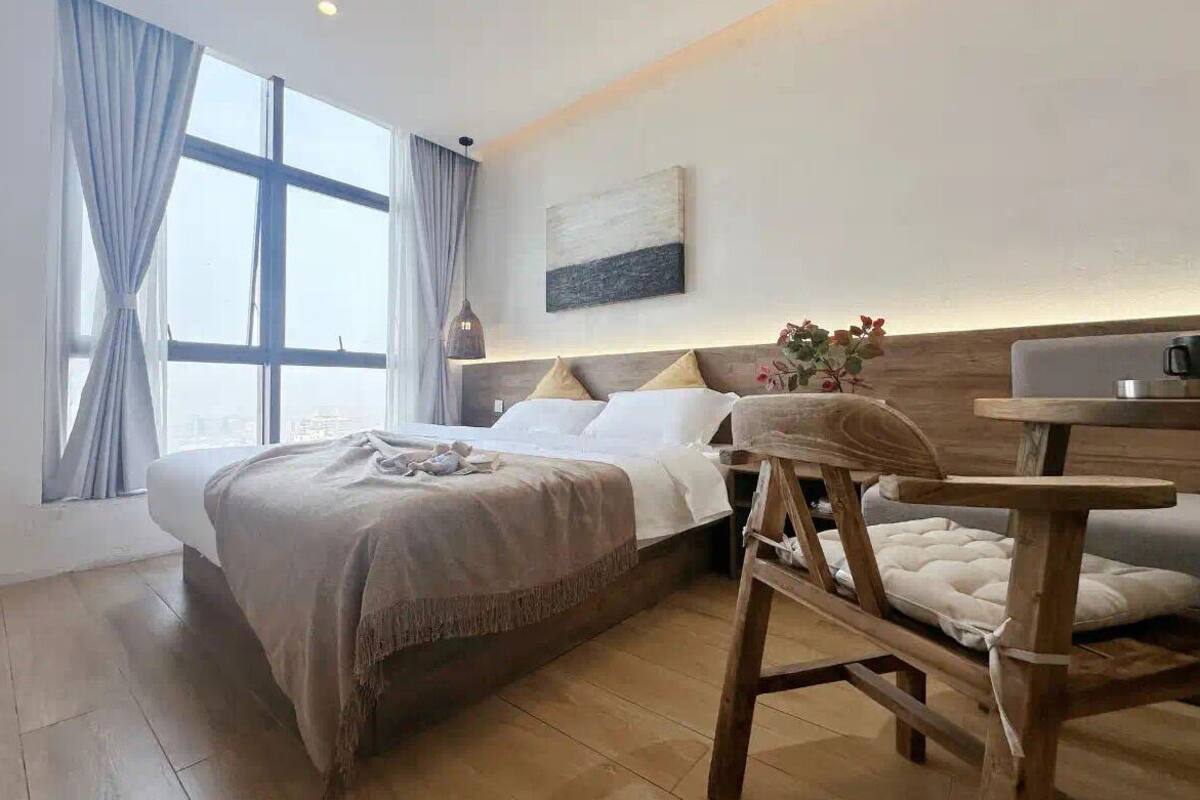 Una camera d'albergo moderna con un letto grande, un comodino in legno, un tavolino e vista sulla città attraverso ampie finestre.